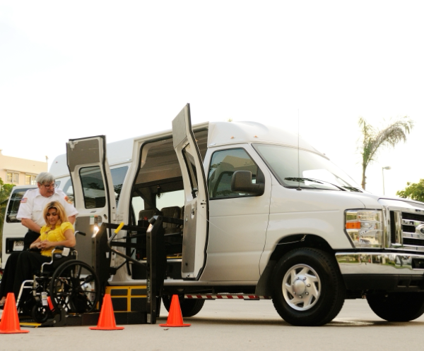 A transportation van with a wheelchair user and assistant using the ramp outdoors, ensuring accessibility.