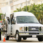 A parking assistant operating a van with a wheelchair lift in a palm-lined parking area.