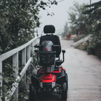 A red mobility scooter parked on a rainy village street path lined with trees and railings.